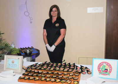 A woman in a uniform stands behind a catering table filled with arranged appetizers at an indoor event.