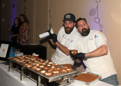 Two chefs in white uniforms preparing food at a banquet, one using a blow torch on desserts.