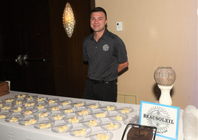 Man in a polo shirt smiling next to a table with trays of cupcakes at a beausoleil restaurant event.