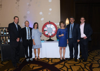 Five adults standing beside a prize wheel and a table with wine bottles at a formal event.