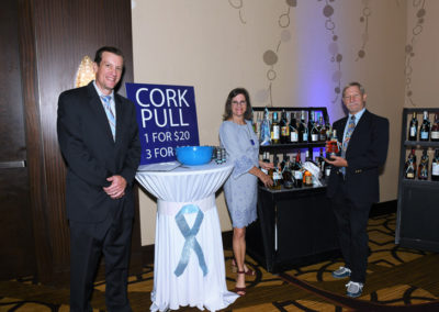 Three people stand around a table labeled "cork pull 1 for $20, 3 for $50" at a charity event, surrounded by bottles of wine.