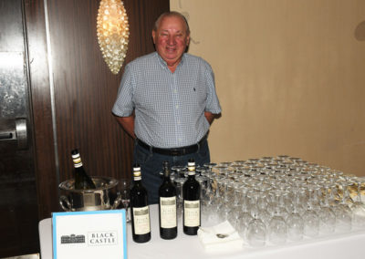 An older man smiling beside a table with wine bottles and glasses at a black castle event.