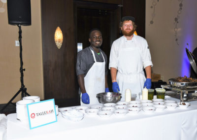 Two chefs, one black and one white, standing behind a food station with prepared dishes at an event, smiling at the camera.