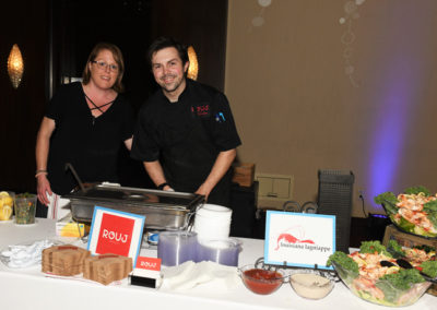 Two caterers stand behind a food station with a sign that reads "rouj" at an indoor event, serving shrimp dishes.
