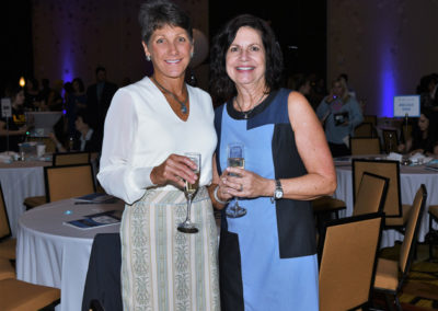 Two women smiling at a formal event, each holding a glass of wine, with banquet tables in the background.
