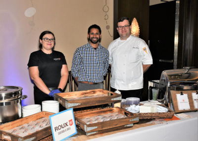 Three chefs standing behind a food station with a roux 61 catering sign at an indoor event.