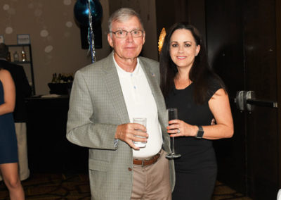 An older man and a younger woman holding drinks at a formal event, standing together with a balloon in the background.