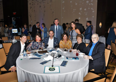 Group of people smiling around a banquet table at an event, with other guests standing and conversing in the background.