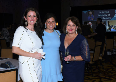 Three women holding drinks and smiling at a gala event, with a tribute presentation visible on a screen in the background.