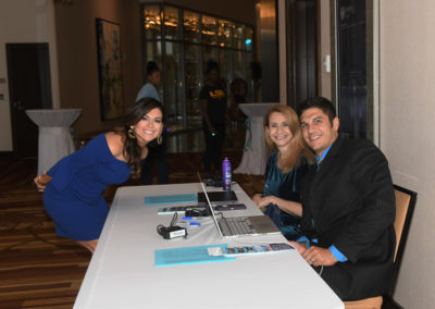 Three smiling people sitting at a registration table with laptops and papers in a conference hall.