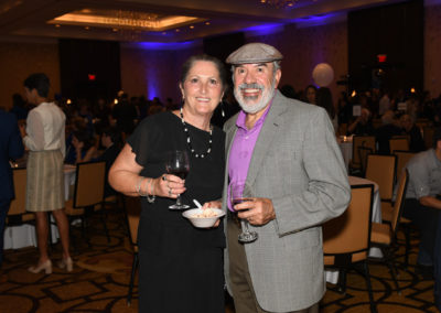 A smiling middle-aged couple holding wine glasses at a social event, with guests and balloons in the background.