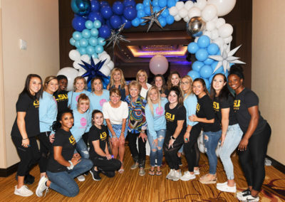 Group of smiling women posing with jill biden under a balloon arch featuring blue and white balloons, wearing casual attire and lsu t-shirts.