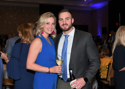 A woman in a blue dress and a man in a suit holding drinks at a social event, smiling at the camera.