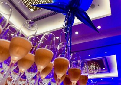 Elegant event setting with a blue balloon sculpture towering over rows of champagne glasses, under a ceiling adorned with twinkling lights. .