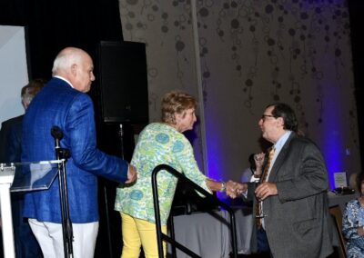 An elderly woman shakes hands with a man at a conference as another man watches from a podium.