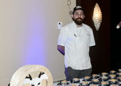 A smiling chef standing beside a cheese wheel and small serving dishes on a table at a catering event.