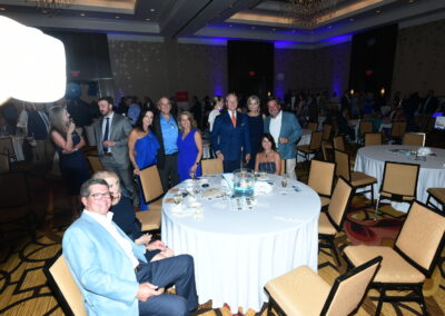 People at a gala event dressed in formal attire, smiling and conversing around tables in a large, dimly lit ballroom with blue lighting.