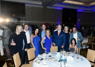 Group of formally dressed adults smiling at a gala event with tables, wine glasses, and a softly lit blue-themed banquet hall background.