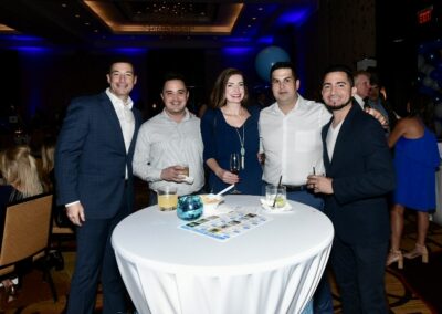 Five people stand smiling around a cocktail table with drinks at a formal event with blue balloon decorations.