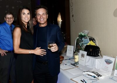 A man and a woman smiling and holding drinks at a charity auction event, standing next to a table with displayed auction items.