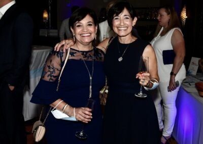 Two women smiling at a party, one holding a wine glass. they wear elegant dresses and stand in a dimly lit room with festive lights.