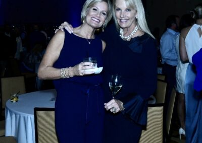 Two women smiling and holding drinks at a social event, dressed in elegant dark dresses.
