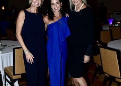 Three women in elegant evening wear smiling for a photo at a formal event, two in black and one in blue, in a dimly lit room with tables.