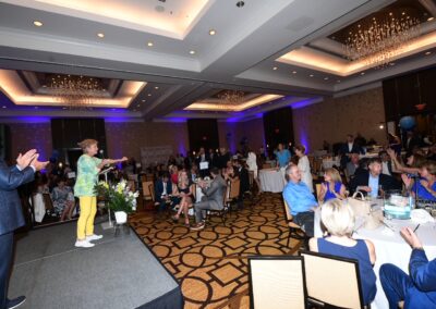 A presenter in colorful clothing speaks to an audience at a well-attended indoor event in a ballroom with chandeliers.