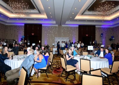 People attending a formal event in a large banquet hall with round tables, chandeliers, and a presentation stage.