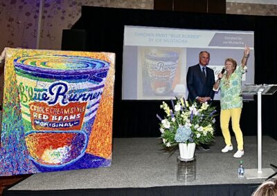 A large, colorful painting of a blue runner red beans can is displayed on stage at an event, with two speakers standing beside a projection screen showing the same image.