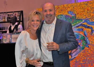 A smiling couple posing in front of a colorful abstract painting at an event, with a bartender in the background.