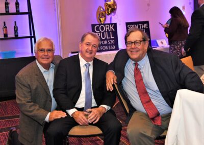 Three smiling men in business attire sitting together at a charity event with "cork pull" and "spin the bottle" signs in the background.