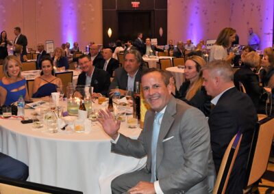 A man in a gray suit smiling and waving at the camera during a formal dinner event with other attendees seated around tables in a well-lit banquet hall.