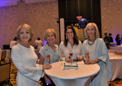 Four women smiling at a social event, holding drinks and standing around a high round table with party decorations in the background.