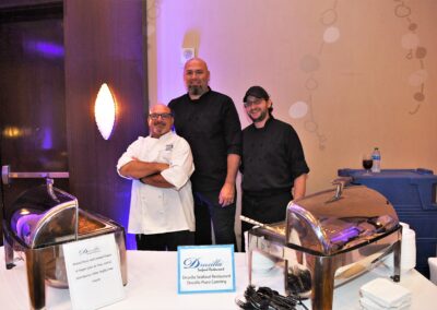 Three chefs, each wearing different styles of black and white chef attire, stand behind serving counters with food warmers, in a dimly lit banquet hall.