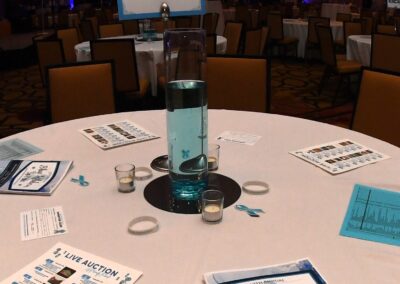 Banquet hall set for an event with tables displaying brochures, a centerpiece with a blue liquid, and a projector screen in the background.