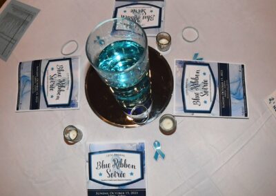A table set for an event with blue ribbon service pamphlets, teal napkins, a centerpiece with a blue candle, and scattered small ribbons.