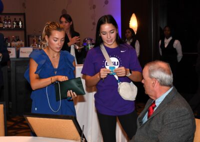Two young women showing documents to an older man sitting at a table during an indoor event.