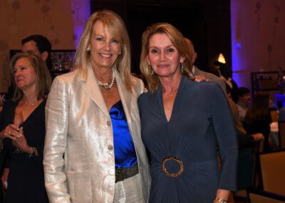 Two smiling women standing together at an event, one in a silver blazer and blue top, the other in a dark blue dress with a circular belt.