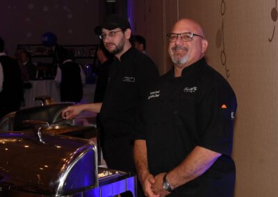 Two male chefs in black uniforms standing by a buffet station inside a well-lit event hall.