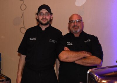 Two chefs, one younger and one older, dressed in black uniforms with embroidered names, posing together with arms crossed in a banquet hall.