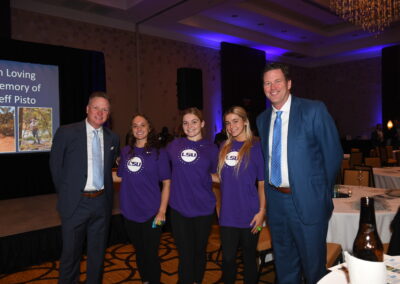 Five people, three in business attire and two in purple lsu shirts, standing together in a banquet hall with a memorial tribute screen in the background.