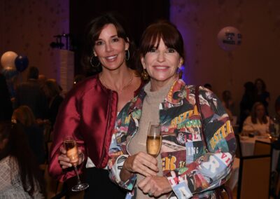 Two women smiling at a party, holding glasses of champagne, with one dressed in a colorful jacket and the other in a satin blouse.