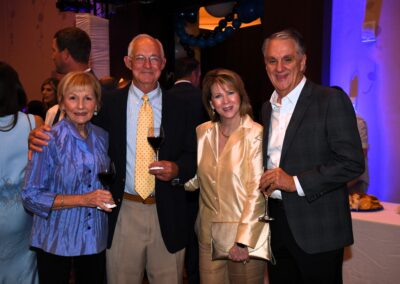 Two senior couples smiling at a formal event, holding wine glasses, with a party atmosphere in the background.