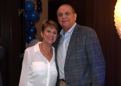 An elderly couple standing together smiling at a party with blue and white balloons in the background.