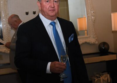 A man in a suit holding a champagne glass at a formal event, standing near a mirror and decorative items.