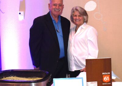 A man and a woman smiling next to a buffet serving station with a sign labeled "roux 61 seafood & grill" at an indoor event.