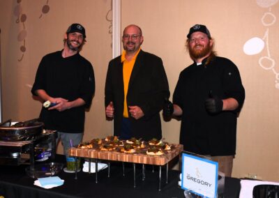 Three men at a catering event, two in chef attire beside a dish display, the other in a suit, all smiling at the camera.