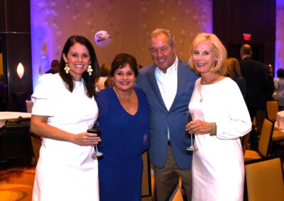 Two women and a man, all smiling, stand with drinks in a banquet hall at a social event. one woman wears a blue dress and the others are in white.