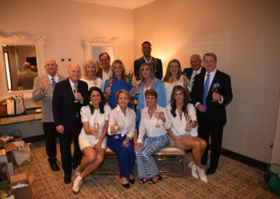 Group of adults in semi-formal attire holding wine glasses and posing for a photo in a well-lit room.